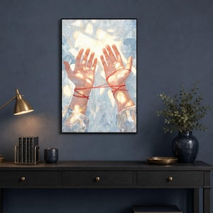 May include: A framed wall art print featuring a painting of two hands reaching upward with thin red strings wrapped around the wrists and palms. The background is a textured, light blue and white abstract wash with bright, glowing highlights.