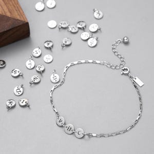 May include: A silver-toned chain bracelet featuring three circular charms engraved with the letters V, B, and C. The bracelet includes a lobster claw clasp and an adjustable extender chain. Several loose, individual circular letter charms are scattered nearby.