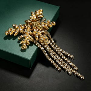 May include: A gold-toned brooch featuring a leaf-inspired design encrusted with clear rhinestones. The piece includes multiple dangling chains of clear rhinestones that extend from the main body, creating a cascading effect of sparkling crystals.