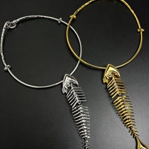 May include: Two circular necklaces, one in silver tone and one in gold tone, each featuring a prominent fish skeleton pendant. The pendants are detailed with small, clear stones set along the spine, suspended from a smooth, rigid metal collar.