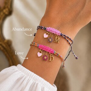 Pode incluir: Duas pulseiras de cordão trançado com seções centrais tecidas em rosa. A pulseira superior tem um coração rosa claro, uma flor roxa e um pingente com a letra M. A pulseira inferior tem um coração rosa claro, uma flor rosa escuro e a letra D.
