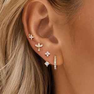 Könnte beinhalten: Ein Set goldfarbener Ohrringe bestehend aus einem sternförmigen Stecker, einem blütenartigen Stecker mit Anhänger, einer vierblättrigen Blume, einem runden Stein-Stecker und einem kleinen Klapp-Creolen-Ohrring mit winzigen klaren Steinen.