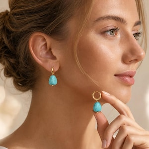 May include: A pair of gold hoop earrings featuring dangling, irregular, teardrop-shaped turquoise stones. The earrings consist of a polished gold circular hoop with a small, vibrant blue-green stone suspended from the bottom of each hoop.