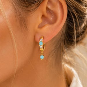 May include: A gold hoop earring featuring two small clear stones and a central turquoise stone on the hoop, with a dangling charm consisting of a single turquoise stone. The earring is secured through the earlobe with a hinged closure mechanism.