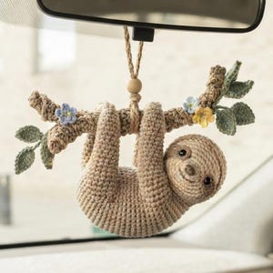 Peut inclure: Un paresseux crocheté beige suspendu à une branche brune texturée, retenu par une ficelle avec une perle en bois. La branche est ornée de petites feuilles vertes et de minuscules fleurs bleues et jaunes, conçu comme un ornement de rétroviseur.
