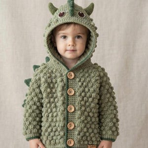 May include: A sage green crocheted hooded cardigan featuring a textured bobble stitch pattern, a hood with small triangular spikes, and five light brown wooden buttons down the front. The garment has long sleeves and a small rectangular label.