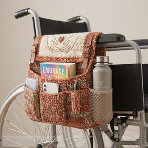 Puede incluir: Un bolso lateral de ganchillo para silla de ruedas en tonos naranja, marrón y crema con correas de piel sintética marrón. La solapa presenta un corazón bordado sostenido por manos. Incluye bolsillos con un libro titulado EMBRACE THE DAY, un teléfono y una botella de agua gris con el texto WELLBEING.