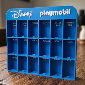 Puede incluir: Un expositor de plástico azul con dieciocho compartimentos rectangulares. La parte superior presenta el texto blanco Disney y playmobil. Cada compartimento está etiquetado con nombres de personajes como Mickey Mouse, Donald Duck, Pinocchio y Peter Pan.