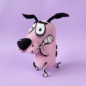 Può includere: Un peluche all'uncinetto a forma di cane realizzato in filato rosa chiaro, con grandi occhi bianchi con pupille nere, un naso nero prominente, orecchie nere e arti neri. Il cane presenta un ampio sorriso con denti e piccole macchie nere sul muso.