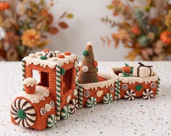 May include: A crochet gingerbread train set featuring a locomotive, a middle car carrying a cone-shaped tree, and a rear car holding a small gift box. The train is decorated with white icing-style trim, peppermint wheels, and candy accents.
