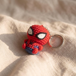 May include: A small, round, crocheted Spider-Man figure keychain. The character features a red head with large white eyes outlined in black, a red and blue body with a small black spider emblem on the chest, and a silver metal keyring attached.