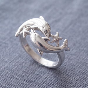 Può includere: Un anello in argento lucido raffigurante due delfini che nuotano in formazione circolare, con una piccola stella marina a cinque punte posizionata tra i loro corpi. Il metallo ha una finitura liscia e riflettente che cattura la luce.