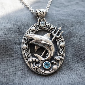 May include: A silver pendant necklace featuring an oval frame decorated with seashells and starfish. Inside the frame, a sculpted shark swims above stylized waves, accompanied by a trident and two small, round, light blue gemstones.