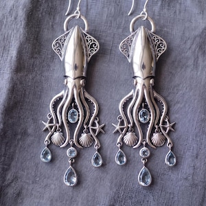 May include: A pair of silver-toned earrings shaped like squids, featuring intricate filigree details on the mantle, dangling starfish and seashell charms, and faceted light blue teardrop and oval gemstones suspended from the tentacles.