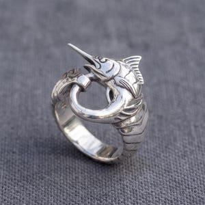 May include: A polished silver ring featuring a detailed marlin fish design. The fish is sculpted to wrap around the finger, with its long, pointed bill curving back toward its tail, creating a circular band shape with visible fin and scale textures.