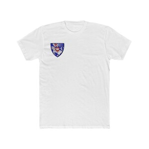 Puede incluir: Una camiseta blanca de manga corta con un pequeño emblema en forma de escudo en el pecho izquierdo. El emblema muestra un fondo azul con una figura de caballo blanco y un detalle de cresta roja en la parte superior, en estilo heráldico.