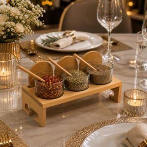 May include: A light wood condiment server featuring three clear glass bowls filled with red pepper flakes, dried green herbs, and ground brown spices. Each bowl includes a small wooden spoon and a circular wooden lid resting behind it.