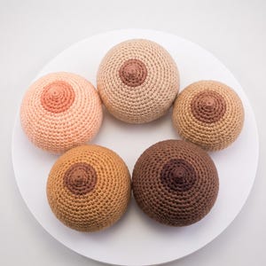 May include: Five spherical, crocheted objects in varying skin-tone shades, each featuring a textured, raised center point. The yarn colors range from light peach and beige to tan, medium brown, and dark brown, arranged in a circular formation.