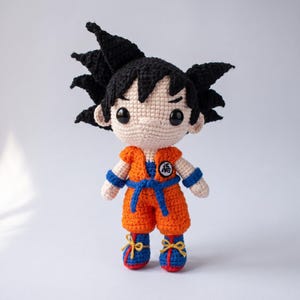 Puede incluir: Una muñeca de ganchillo que representa un personaje con pelo negro puntiagudo, grandes ojos negros y piel clara. La muñeca viste un gi naranja con cinturón azul, muñequeras azules y botas azules con cordones amarillos. En el pecho lleva un parche circular blanco con el kanji negro 悟.
