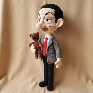 May include: A handmade crochet doll depicting a character with black hair, a beige face, and a prominent nose. The doll wears a grey jacket, white shirt, red tie, and black trousers. It holds a small brown crochet teddy bear in its right hand.