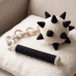 May include: A crocheted flail toy featuring a cream-colored spherical head with black spikes, a cream-colored chain, and a black cylindrical handle. The entire object is crafted from yarn with a visible stitch texture, showcasing a two-tone color scheme.