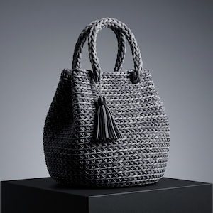 Puede incluir: Un bolso de mano de ganchillo en color gris oscuro con un patrón tejido texturizado, dos asas trenzadas y una borla negra decorativa sujeta a una cadena plateada. El bolso tiene una silueta redondeada tipo cubo con tejido uniforme.