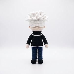May include: A handcrafted crochet doll featuring a character with spiky white hair and a white blindfold covering the eyes. The figure wears a black long-sleeved sweater with white trim at the collar and cuffs, paired with dark blue trousers and black shoes.