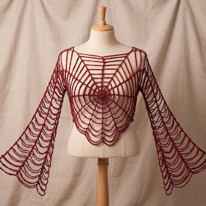 May include: A dark red crochet top featuring a spider web pattern that extends from the center of the chest across the long, flared sleeves. The garment has a wide neckline and a cropped, pointed hemline, constructed with openwork yarn loops.