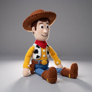 Op de afbeelding: Een gehaakte pop van een cowboyfiguur met een bruine hoed, een rode halsdoek, een wit vest met zwarte koeienvlekken, een geel shirt met lange mouwen, een blauwe broek, een bruine riem met een gouden gesp en bruine laarzen met gele accenten.