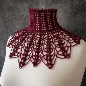 May include: A deep burgundy crochet collar featuring a high neck design with a vertical ribbed pattern and a wide, scalloped lace-like edge that drapes over the shoulders. The intricate needlework creates a delicate, open-work floral motif.