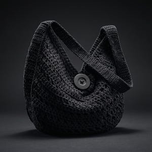 Puede incluir: Un bolso hobo de ganchillo negro con un patrón tejido texturizado, una sola correa de hombro integrada y un botón negro circular grande centrado en la parte delantera. El bolso tiene una forma redondeada y holgada con un acabado oscuro.