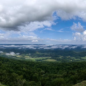 May include: A panoramic view from a high elevation overlooking a vast landscape of dense green forests and rolling hills under a bright blue sky filled with scattered white clouds and a low-hanging layer of mist stretching across the horizon.