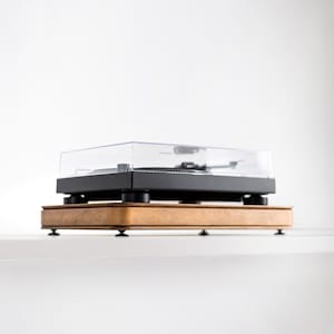 Op de afbeelding: Een zwarte platenspeler met een doorzichtige plastic stofkap staat op een rechthoekig houten isolatieplatform. Het platform heeft een warme bruine houtnerfafwerking en wordt ondersteund door vier kleine, donkere, ronde voetjes aan de onderkant.