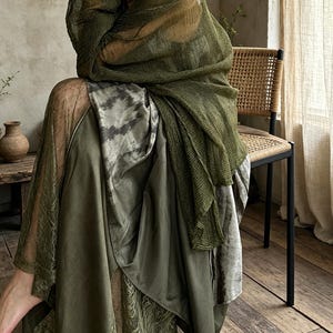 Puede incluir: Un conjunto de capas que presenta un chal de tejido abierto en color verde oliva sobre un vestido largo. El vestido consta de una falda verde oliva lisa, un panel con estampado tie-dye en tonos grises y verdes, y detalles de encaje.