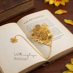 May include: A gold-toned metal bookmark featuring a detailed sunflower bouquet design, connected by a delicate chain to a smaller sunflower charm. The bookmark rests on an open book page with the printed text SUNFLOWER and With angel eyes.