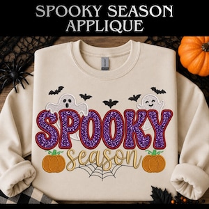 May include: A sand-coloured crewneck sweatshirt featuring an embroidered design with the purple glittery text SPOOKY and the gold cursive text season. The graphic includes two white ghosts, black bats, two orange pumpkins, and a black spiderweb.
