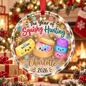 May include: A round, clear, faceted glass ornament featuring a red ribbon hanger. The design includes the text "The Year of Squishy Hunting", "Charlotte", and "2026". Graphics show a stick of salted butter, a dumpling in a steamer, and three stacked squishy toys.