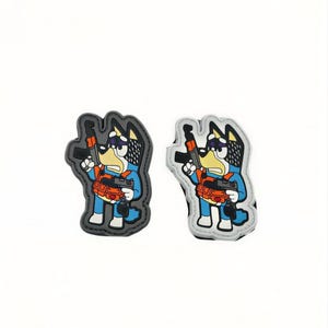 Könnte beinhalten: Zwei gummierte Aufnäher mit einer Zeichentrick-Hundefigur in blauem Outfit, die eine orangefarbene taktische Weste trägt und eine schwarze Schusswaffe hält. Ein Aufnäher hat einen dunkelgrauen Rand, der andere einen hellgrauen Rand, beide mit genähten Kanten.