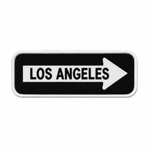 May include: A rectangular embroidered patch with a black background and a white border. The patch features a white horizontal arrow pointing to the right, containing the black text LOS ANGELES in a bold, sans-serif capital letter font.