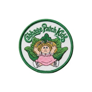 Puede incluir: Un parche bordado circular que presenta el logotipo de Cabbage Patch Kids en texto verde sobre un personaje de dibujos animados con cabello rubio en coletas, vestido con un vestido rosa, sentado frente a grandes hojas verdes sobre fondo blanco.