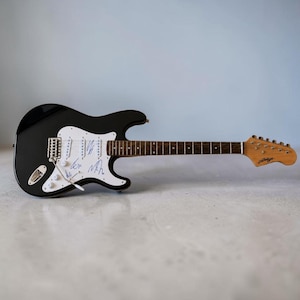 May include: A black electric guitar with a white pickguard, featuring three single-coil pickups, a tremolo bridge, and a wooden fretboard. The white pickguard is decorated with several blue ink signatures. The headstock is light brown wood.