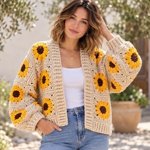 May include: A beige crochet cardigan featuring a repeating sunflower motif with yellow petals and dark brown centres. The garment has long sleeves, a ribbed trim along the front opening, and a loose fit, worn over a plain white top and blue jeans.