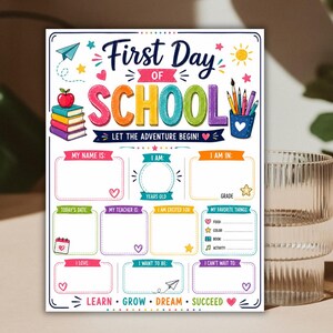 Op de afbeelding: Een wit bord voor de eerste schooldag met kleurrijke tekst, een stapel boeken en een pennenhouder. Het sjabloon bevat invulbare secties voor naam, leeftijd, leerjaar, datum, leraar, favorieten en doelen, versierd met sterren en hartjes.
