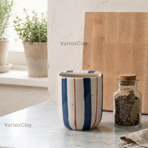 May include: A cylindrical ceramic utensil holder featuring a white base with vertical navy blue stripes and thin red pinstripes. The surface has a speckled texture. The text VortexClay is visible on the side of the vessel and in the background.
