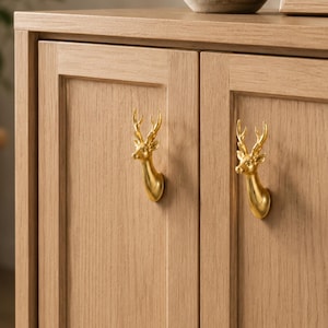 May include: A light oak wooden cabinet featuring two doors, each equipped with a polished gold-colored metal deer head handle. The handles are shaped like the head and antlers of a stag, providing a functional grip for opening the cabinet doors.