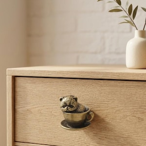 May include: A brass-toned metal drawer knob shaped like a cat sitting inside a teacup with a saucer base. The knob is attached to a light-colored wooden drawer front, featuring a detailed sculpted feline face and a rounded cup handle design.