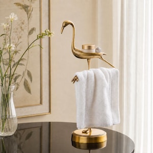 May include: A gold-toned metal flamingo-shaped towel holder featuring a circular base and a small flat platform on the bird's back. A white rectangular towel is draped over the flamingo's legs, and a small round container sits on the bird's back.