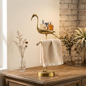 May include: A gold-toned metal bird-shaped stand featuring a flat platform on its back for holding small items and a horizontal bar below for hanging a towel. The stand has a circular base and holds a beige waffle-textured hand towel.