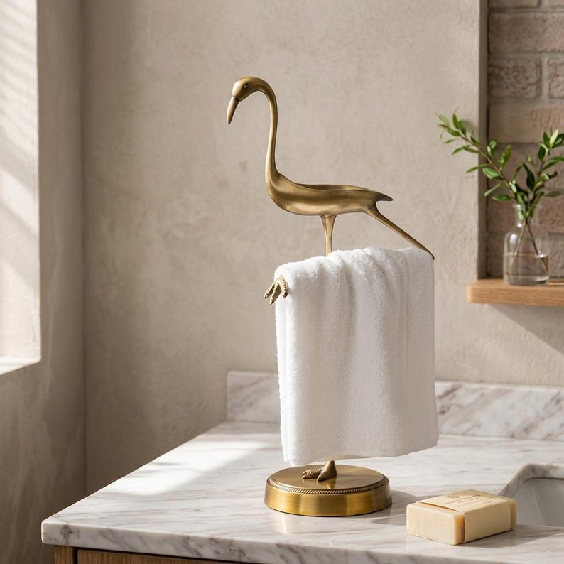 May include: A metallic gold-toned bird-shaped towel holder featuring a long neck and slender legs, standing on a circular base. A plain white rectangular towel is draped over the bird's legs, which serve as a horizontal bar for hanging.