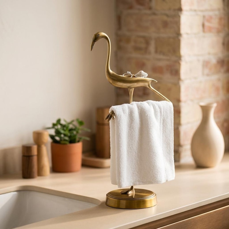 May include: A brass-colored metal bird-shaped stand with a circular base, featuring a flat surface on the bird's back for small items and a horizontal bar for hanging a white towel. The bird has a long neck and stands on two feet.